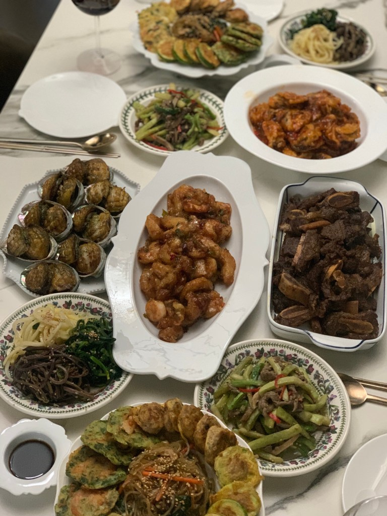 다양한 한식 요리가 담긴 테이블 위의 음식. 잡채, 튀김, 불고기, 해물 요리, 샐러드 등이 포함되어 있으며, 옆에 와인 한 잔이 놓여있다.