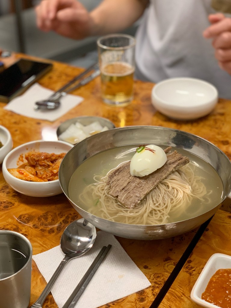 A bowl of cold noodles topped with a slice of beef and a soft-boiled egg, accompanied by side dishes of kimchi and pickled vegetables on a wooden table.