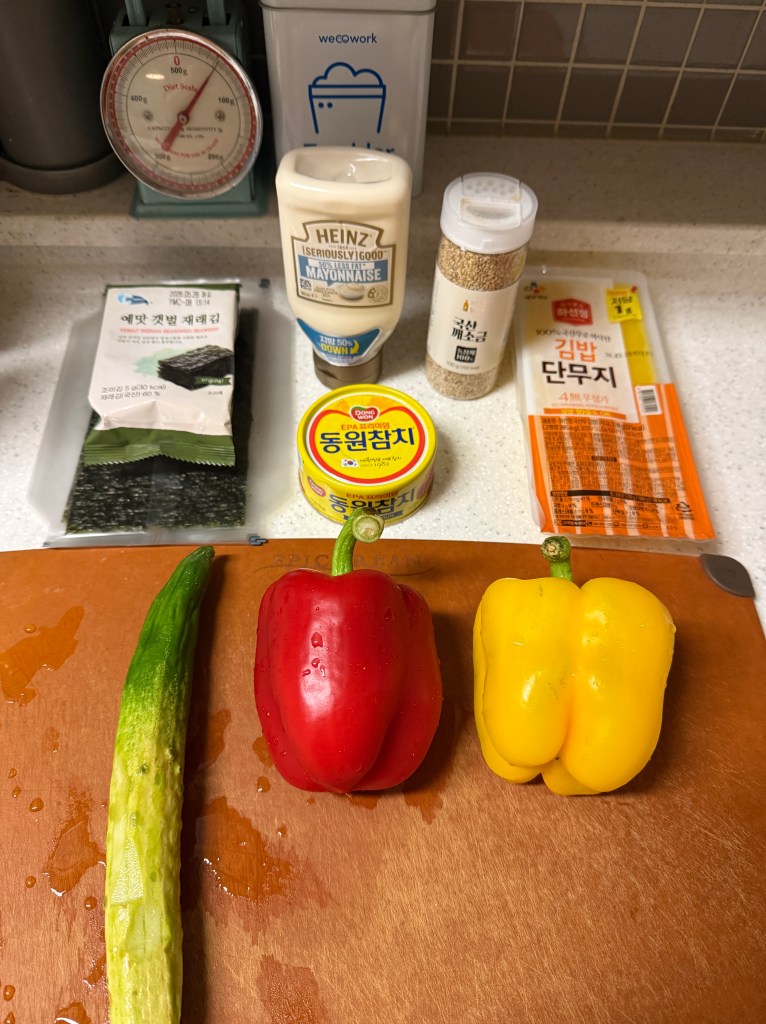A wooden cutting board with a cucumber, a red bell pepper, and a yellow bell pepper arranged in a row. In the background, various food items are displayed, including seaweed, mayonnaise, canned tuna, sesame seeds, and a pack of sushi seaweed.
