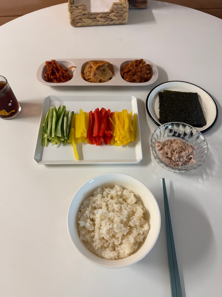 A dining table set with a bowl of rice, colorful sliced vegetables (green onions, yellow and red bell peppers), assorted side dishes in small bowls, a plate of seaweed, and a bowl of tuna.