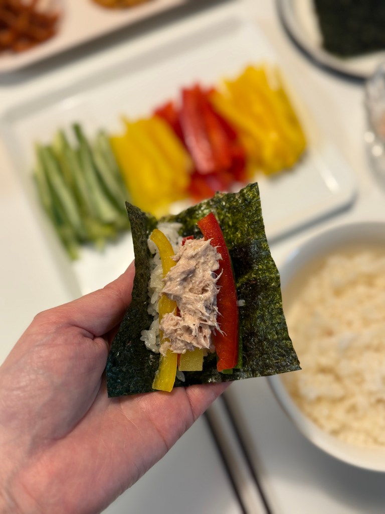 A hand holding a piece of nori wrapped around rice, tuna, and colorful sliced vegetables, including red and yellow bell peppers, in a kitchen setting.