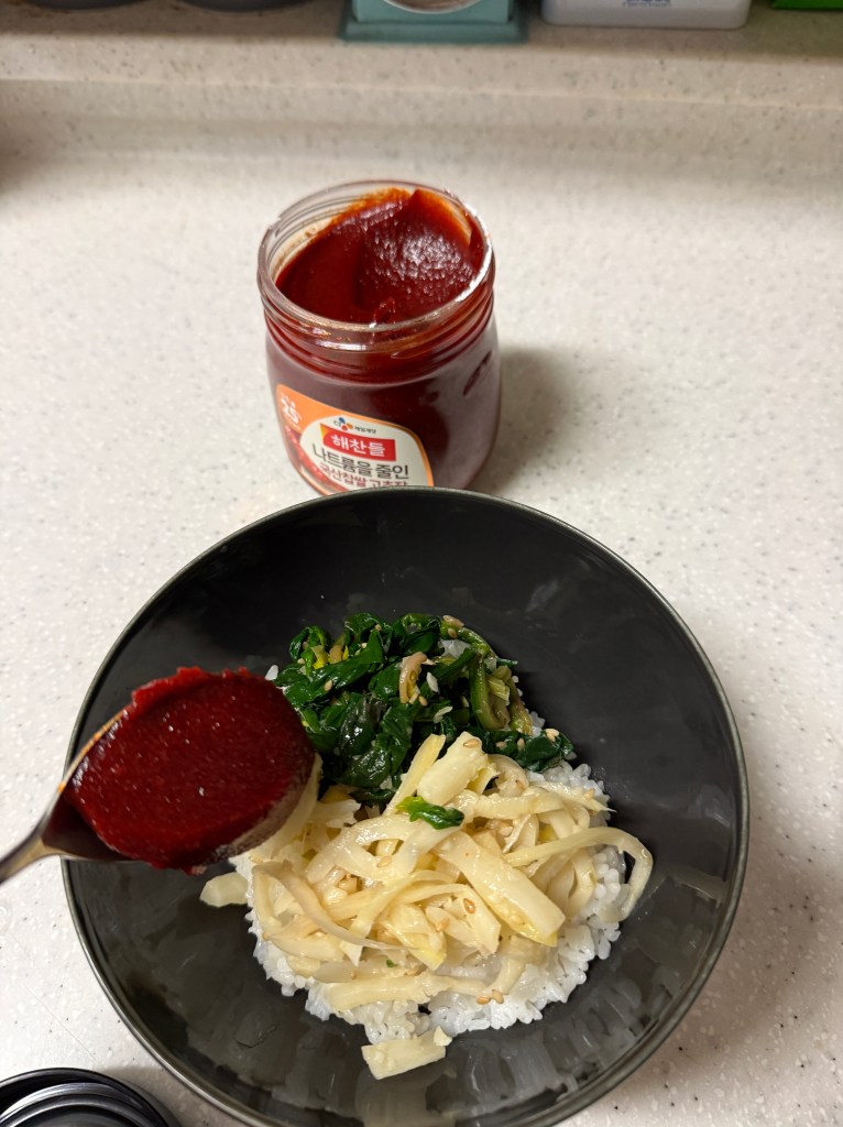 A bowl of rice topped with vegetables, including sautéed greens and bamboo shoots, with a spoonful of red chili paste in the foreground and a jar of the paste in the background.