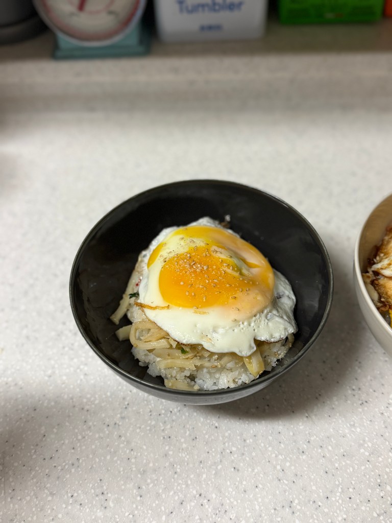 A bowl of rice topped with sautéed vegetables and a fried egg, placed on a countertop.