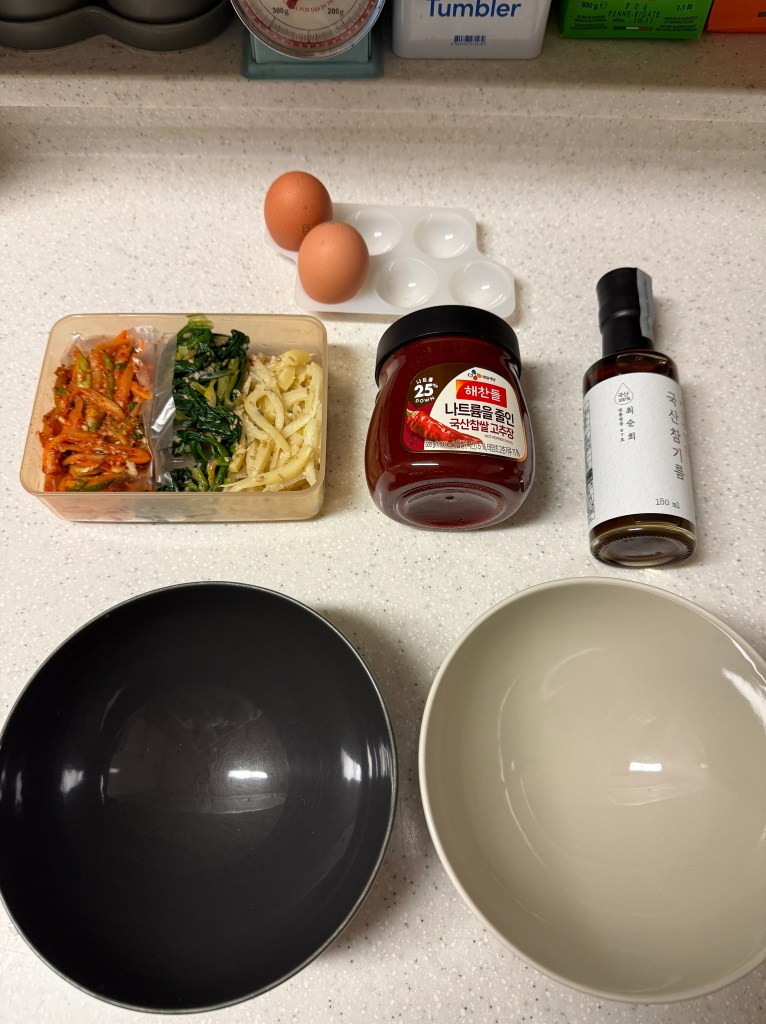 A kitchen countertop with two eggs, a plastic container of assorted vegetables, a jar of Korean chili paste, a small bottle of sauce, and two bowls (one black and one cream-colored).