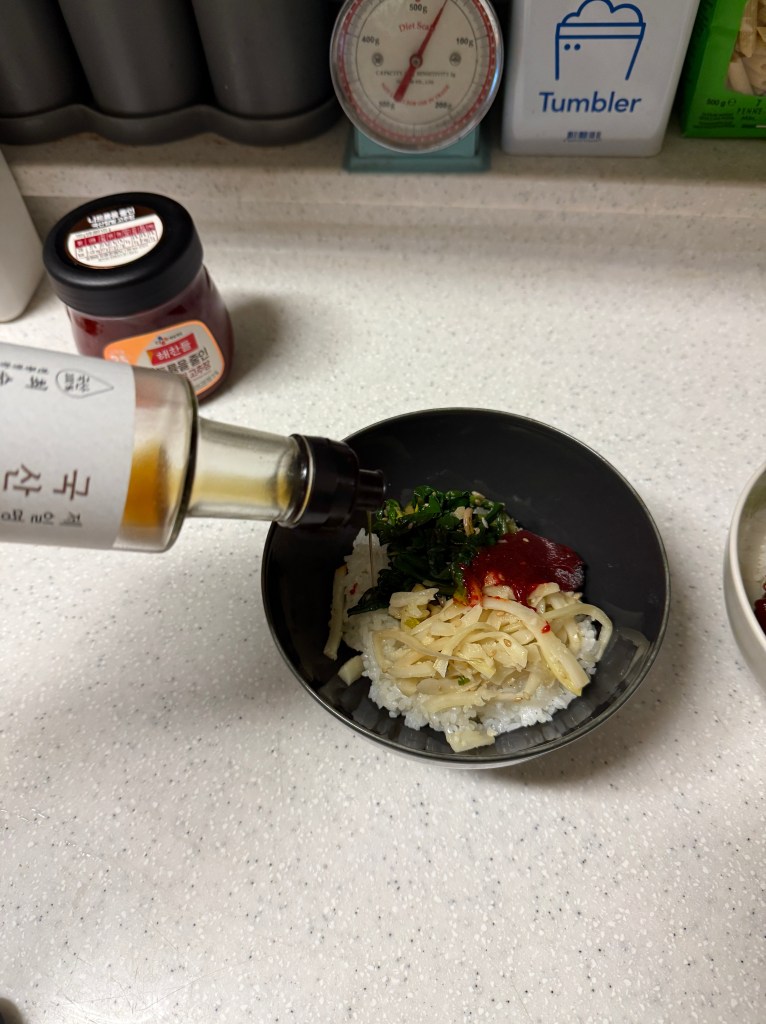 A person is pouring a sauce over a bowl of rice topped with greens and noodles on a countertop.