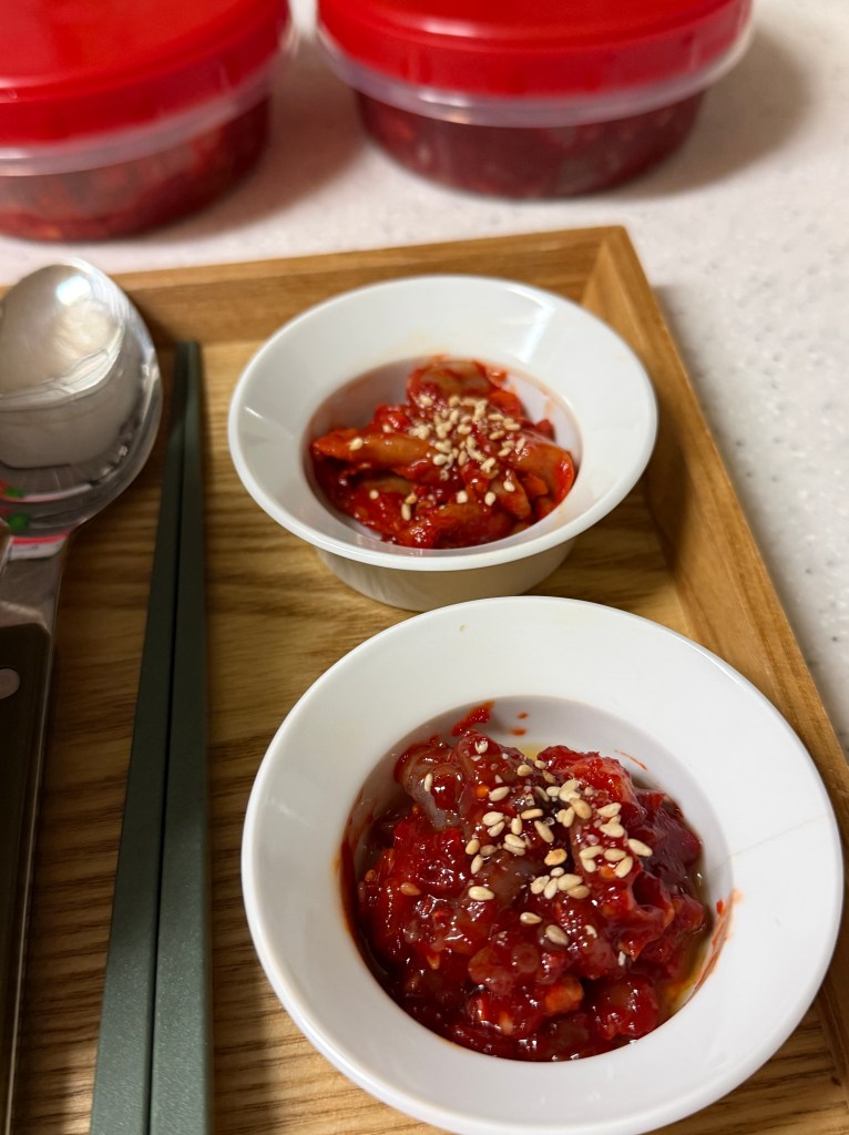 Two small white bowls filled with red spicy sauce topped with sesame seeds, placed on a wooden tray alongside chopsticks and a spoon, with two containers of similar sauce in the background.