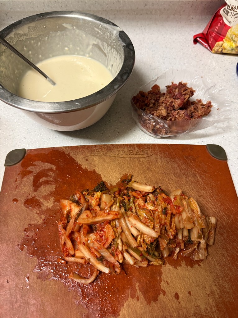 A wooden cutting board with chopped kimchi, a bowl of batter, and a container of meat on a kitchen countertop.