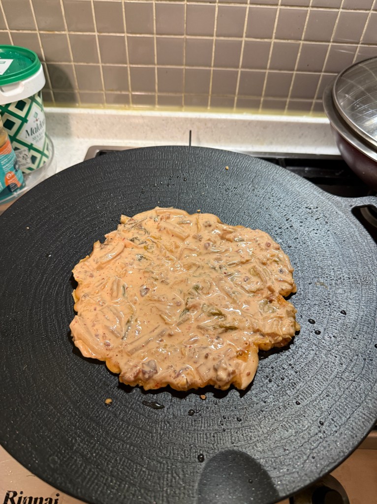 A partially cooked savory pancake filled with vegetables on a black cooking surface.