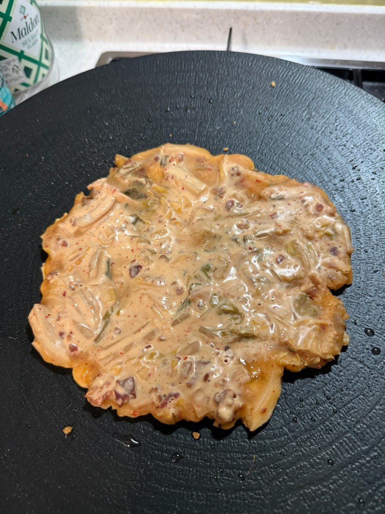 A savory pancake cooking on a black skillet, featuring a mix of vegetables and a creamy batter.