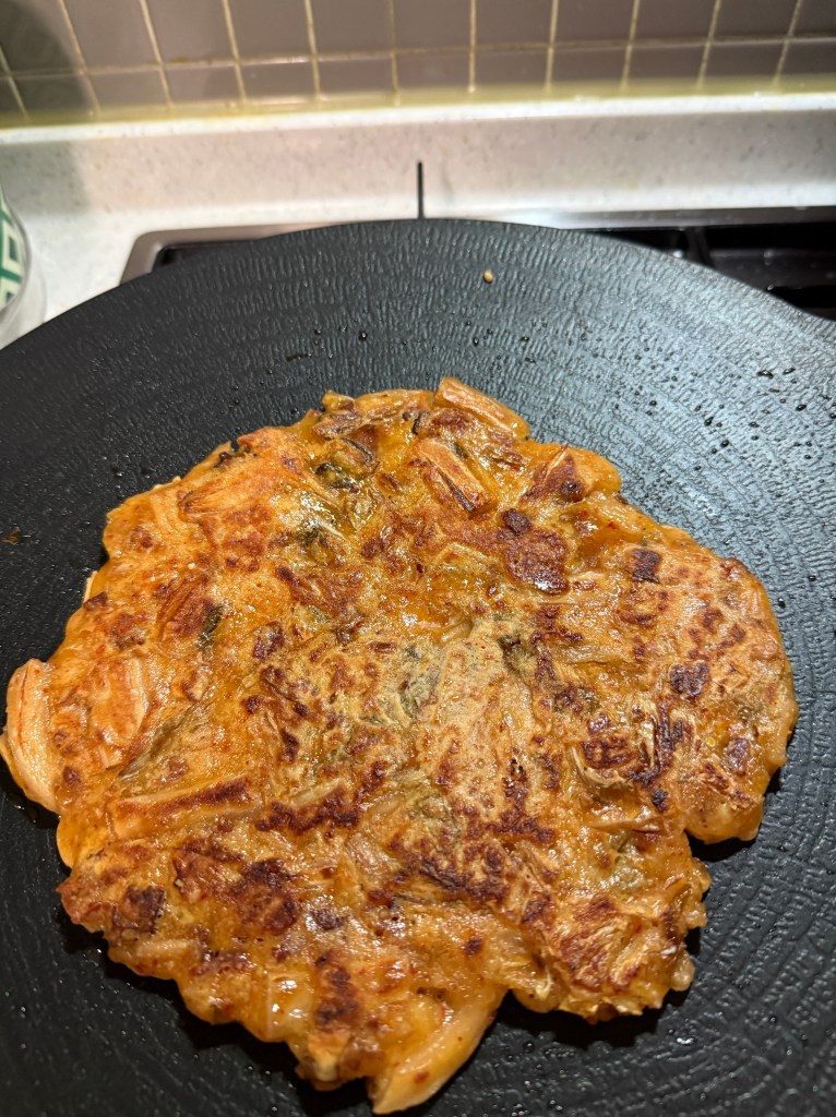 A golden brown savory pancake cooking on a black pan, featuring crispy edges and a textured surface.