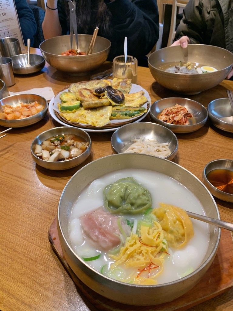 다양한 한국 요리가 담긴 테이블, 중앙에는 만두국이 담긴 그릇이 있으며, 그 주위에는 여러 가지 반찬과 전이 놓여 있다.