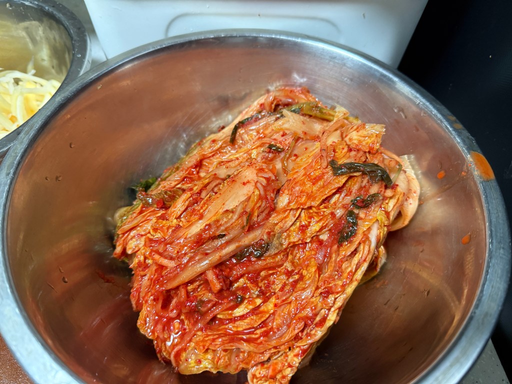 A metal bowl filled with freshly prepared kimchi, featuring vibrant red chili paste and slices of vegetables.