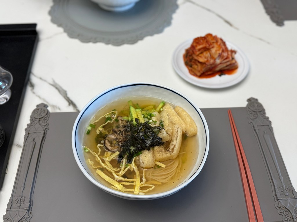 A bowl of clear soup with noodles, topped with green onions, seaweed, and sliced egg. A plate of kimchi is in the background, alongside chopsticks and a placemat.