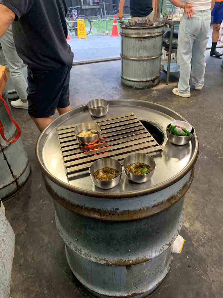 Korean BBQ being grilled on a unique drum can grill in a standing restaurant.