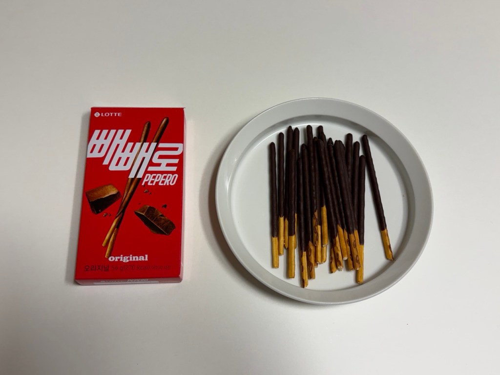 A box of Lotte Pepero snacks featuring chocolate-covered biscuit sticks, alongside a white plate with several Pepero sticks arranged neatly.