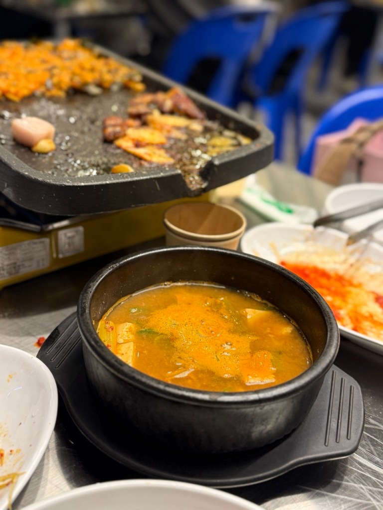 A wide shot showing the stew alongside grilled pork belly on a blue outdoor table.
