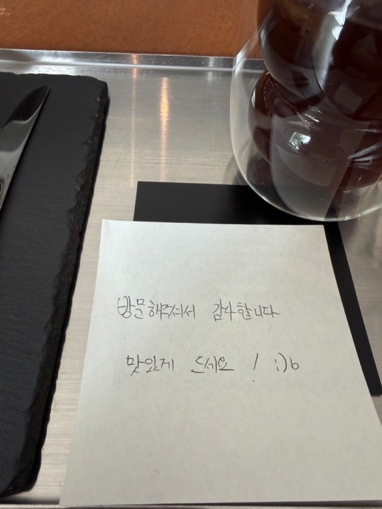 An aesthetic top-down shot of a barista's minimalist thank you letter and coffee flavor note card on a sleek table at Compile Hongdae.