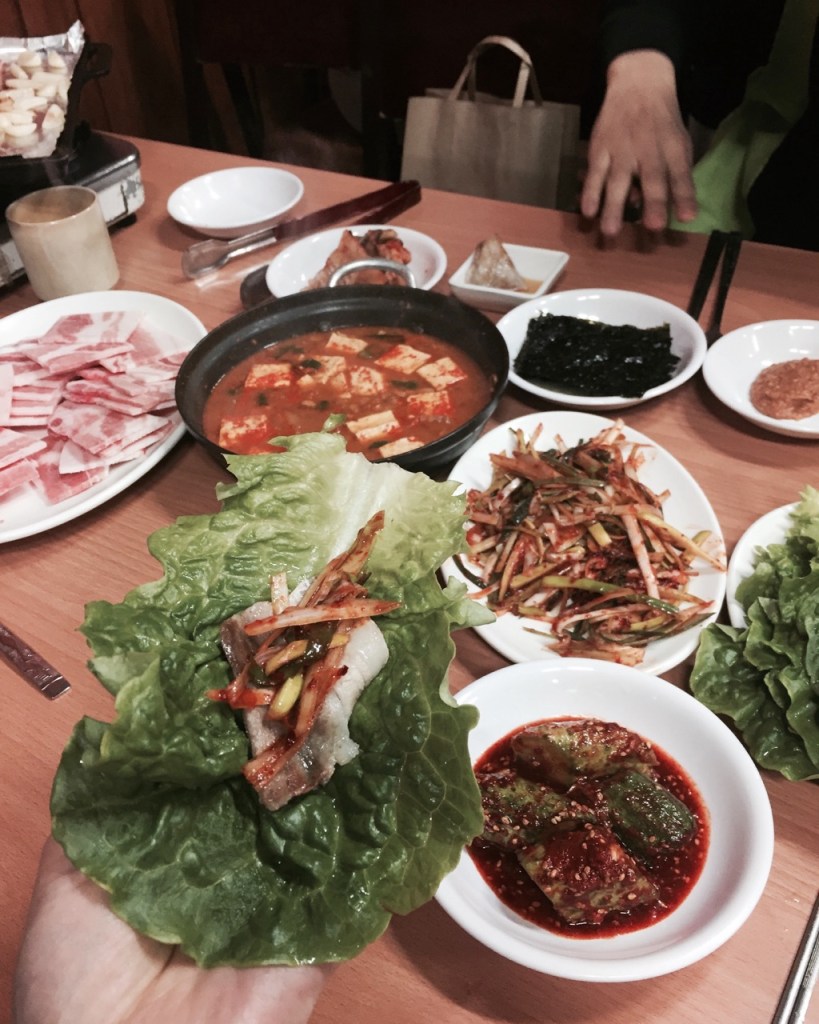 A mouthwatering, close-up shot of a classic Korean pork belly wrap (Ssam) held in hand, loaded with grilled frozen pork, scallion salad, and ssamjang.