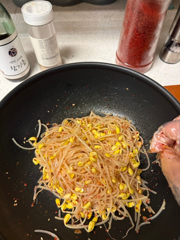 A detailed macro shot showing hands delicately tossing cooked soybean sprouts with toasted sesame oil and seasonings.
