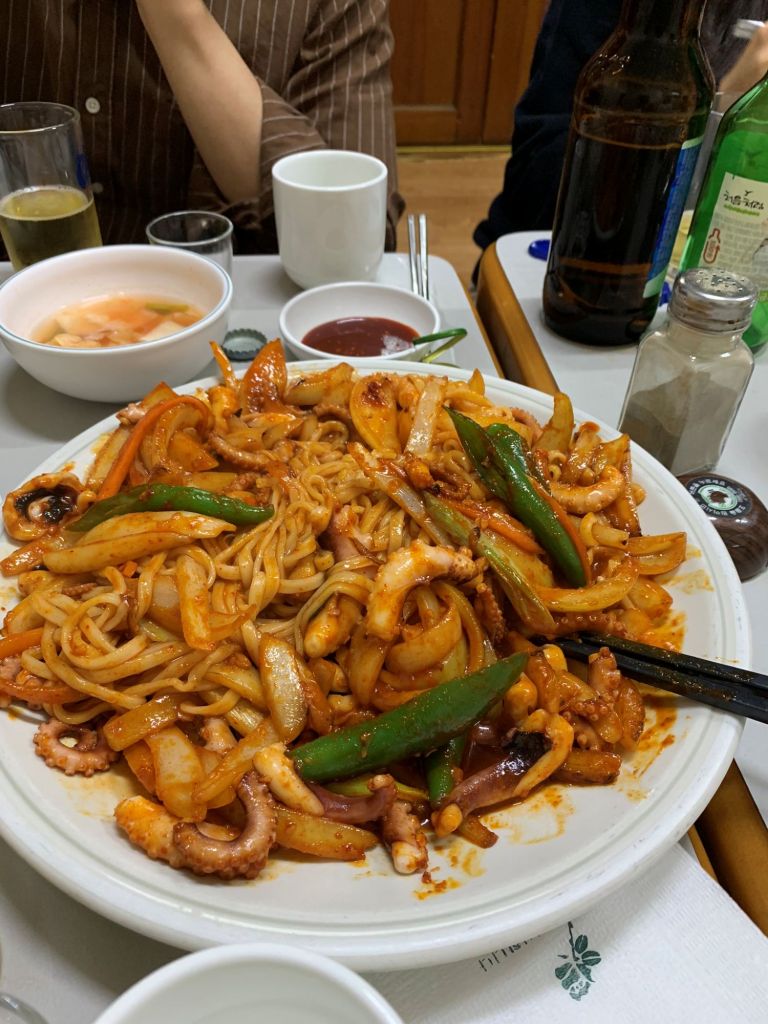 An aesthetic lifestyle shot of chopsticks tossing soft somyeon noodles into the pungent, spicy octopus reduction.