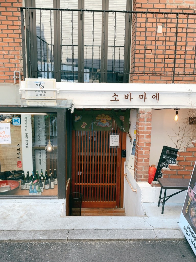 The beautiful and serene entrance facade of Sobamae in Seongsu-dong, featuring a minimalist Japanese wooden door and noren curtain.