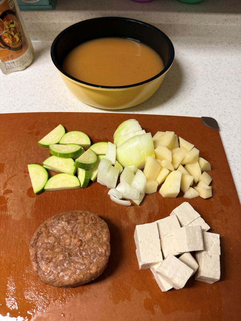 An aesthetic lifestyle snapshot showing prepped ingredients including sliced tofu, radishes, zucchini, and chilies on a wooden board.