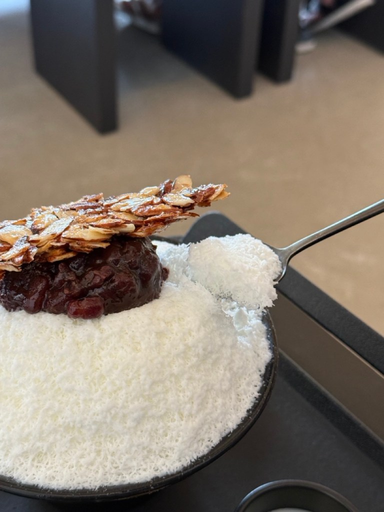 An aesthetic lifestyle snapshot showing a brass spoon scooping up fresh milk shaved ice combined with sweet red beans.