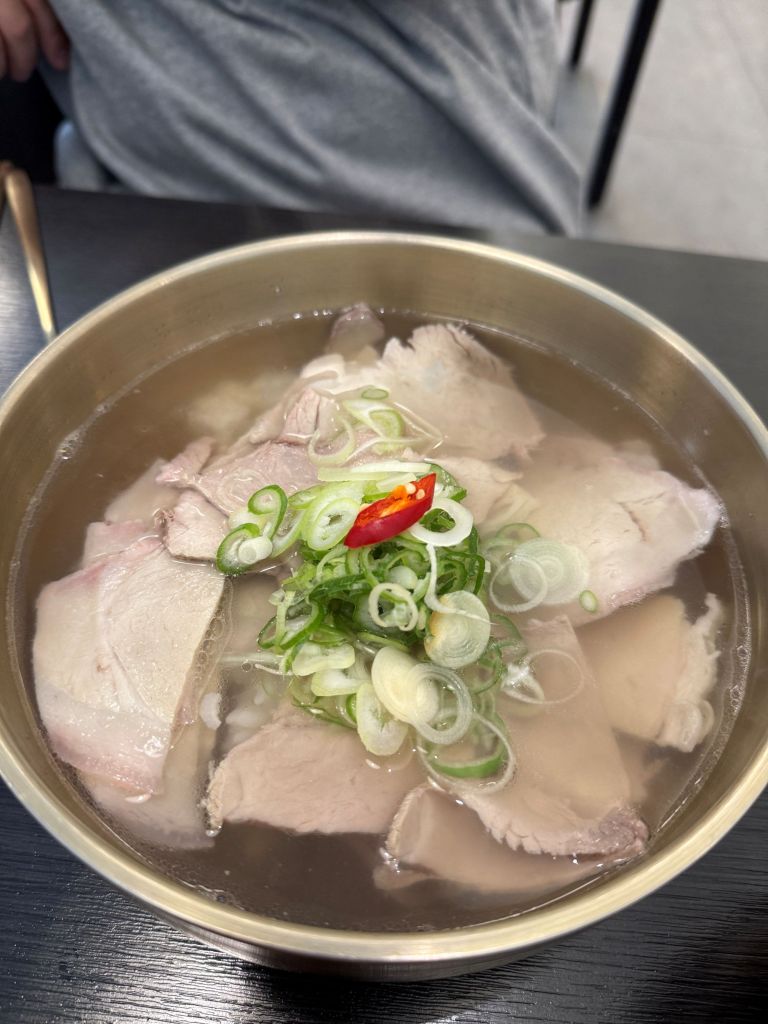 A high-satisfaction top-down view of a steaming brass bowl of clear herbal pork gomtang soup served with side dishes at Dongol Yagbang Seongsu.